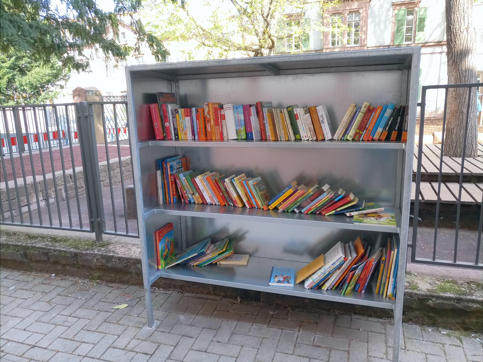 Foto eines Bücherregals im Freien. Es ist etwa zur Hälfte mit Büchern gefüllt.