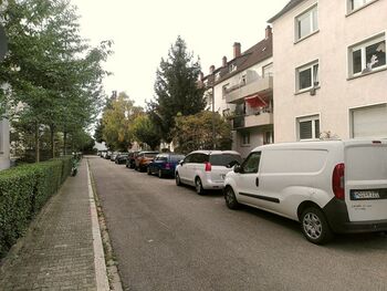 Blick in die Straße. Links ein Gehweg, rechts parken Autos. Es gibt grüne Vorgärten mit hohen Bäumen.